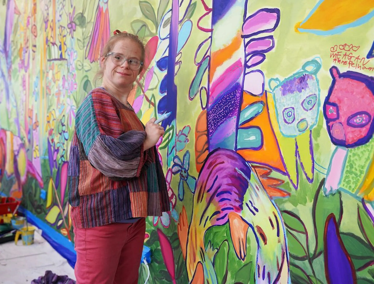 Studio A artist stands in front of a large, vibrant abstract mural painted on a wall. The mural features bright green, purple, pink, orange, and yellow shapes and figures.